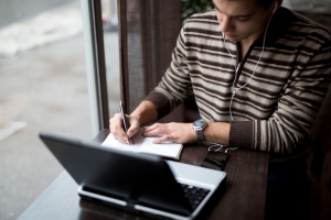 A man takes notes while listening to a presentation on his laptop