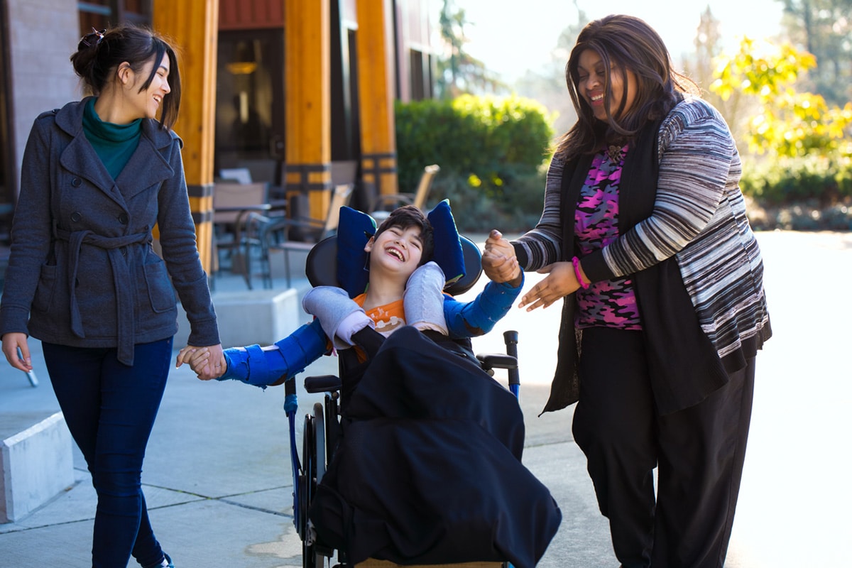 Caregivers hold hands with boy in wheelchair.