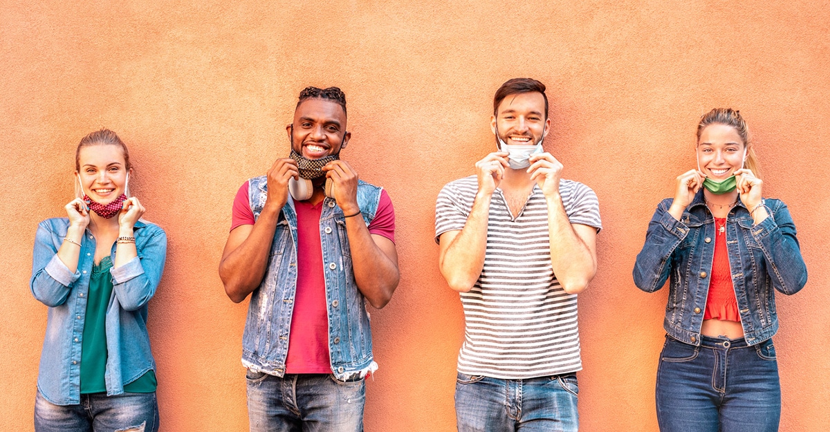 Four people hold their face masks at their chins so the camera can see them smile.
