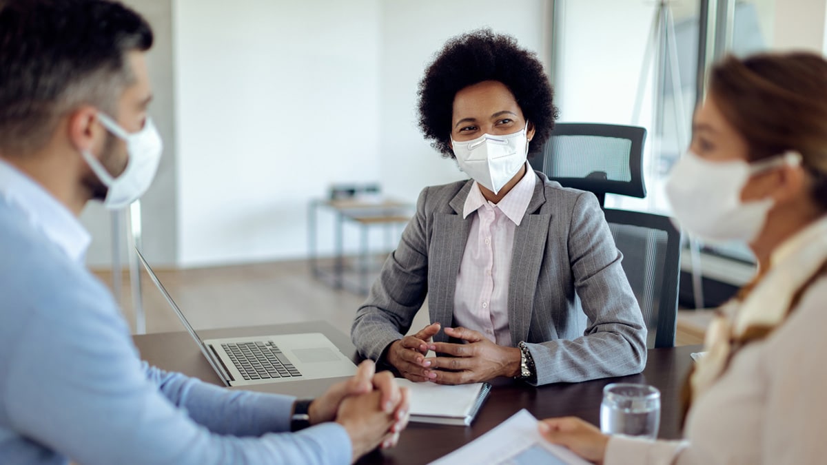 Three people gathered at a desk over paperwork and a laptop, wearing face masks.