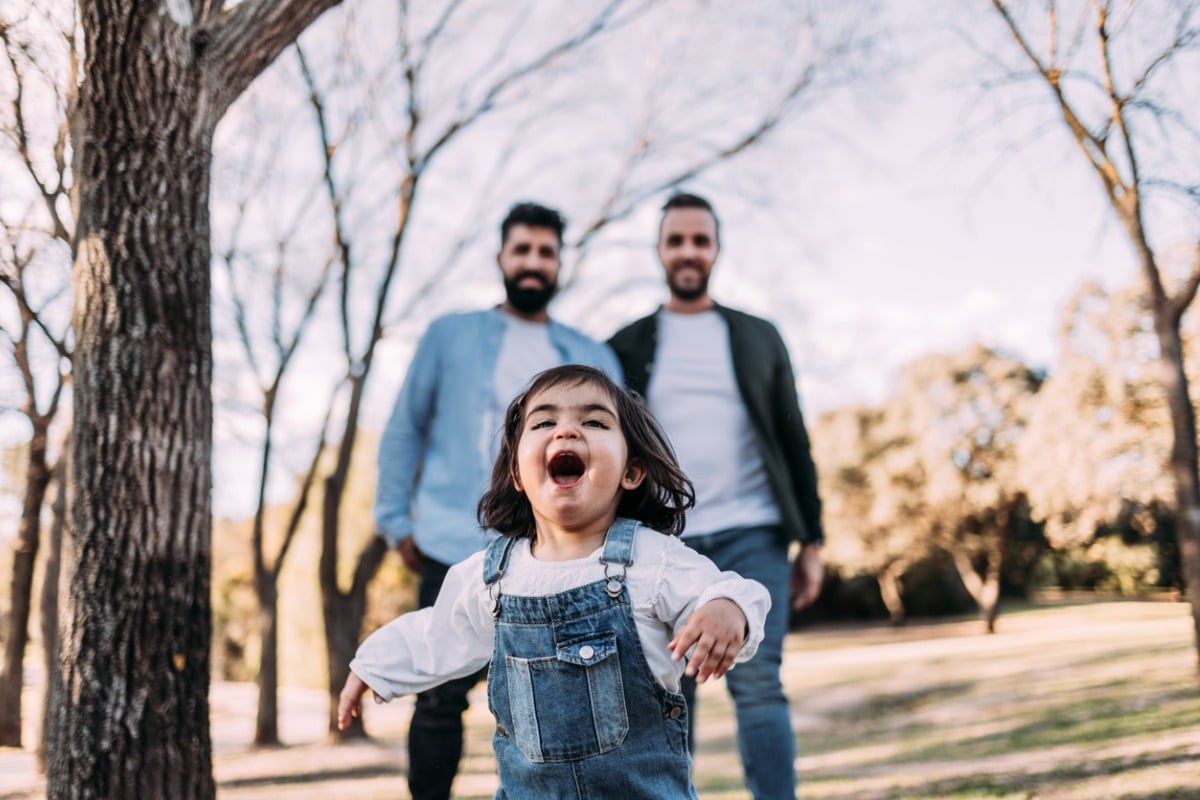 Same sex couple's daughter runs gleefully in front of them in a park.