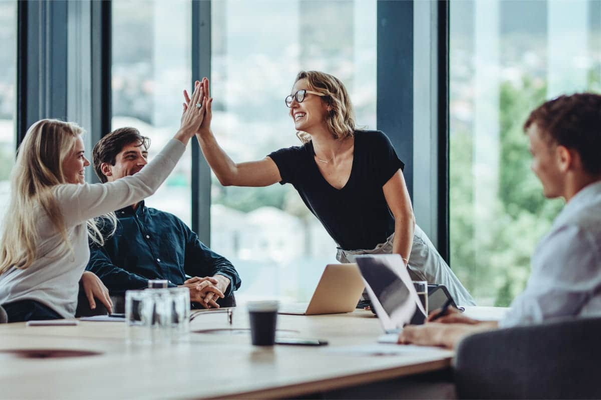group of colleagues sitting around table, two colleagues high-fiving eachother