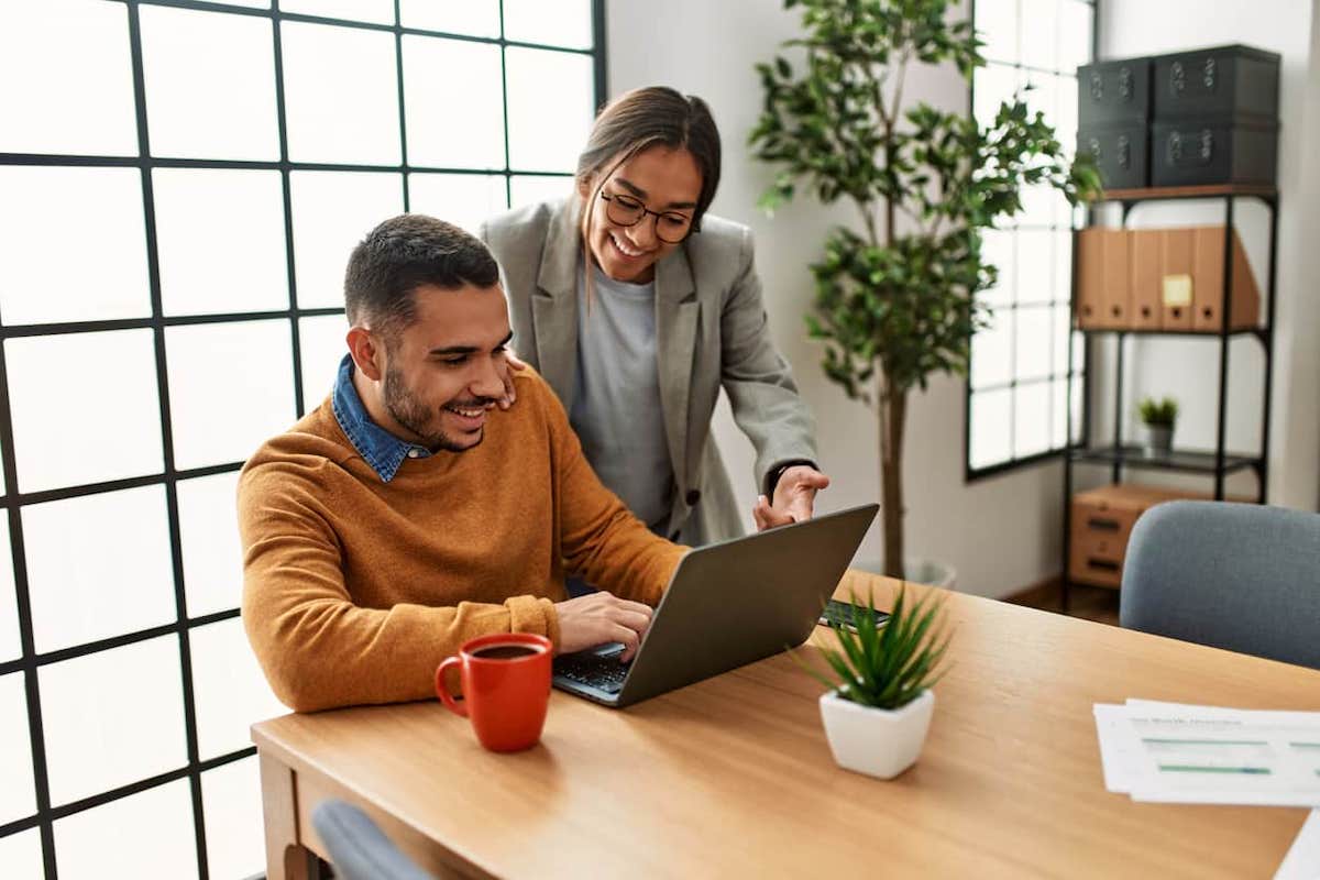 A couple sitting at a desk and working on a laptop. A Spousal Open Work Permit allows spouses of PRs or citizens to work in Canada.