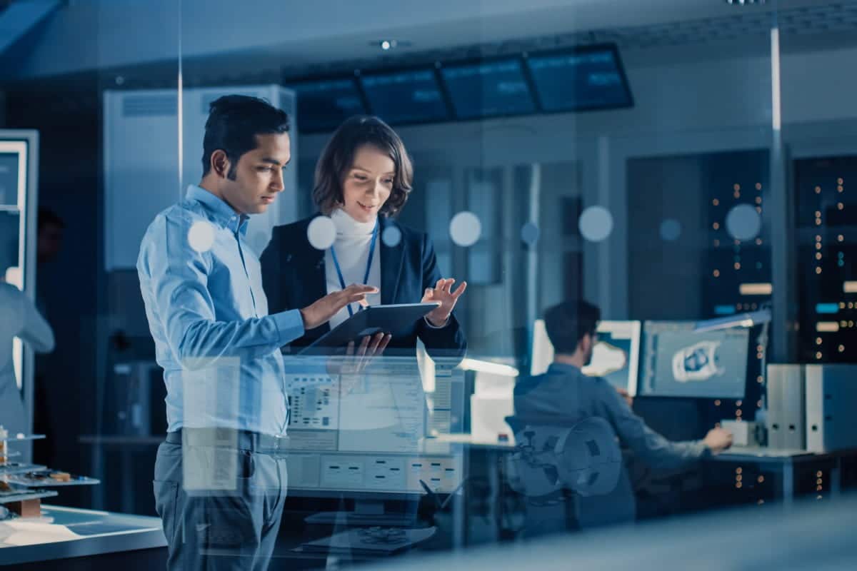 In Technology Research Facility: Female Project Manager Talks With Chief Engineer, they Consult Tablet Computer. Team of Industrial Engineers, Developers Work on Engine Design Using Computers