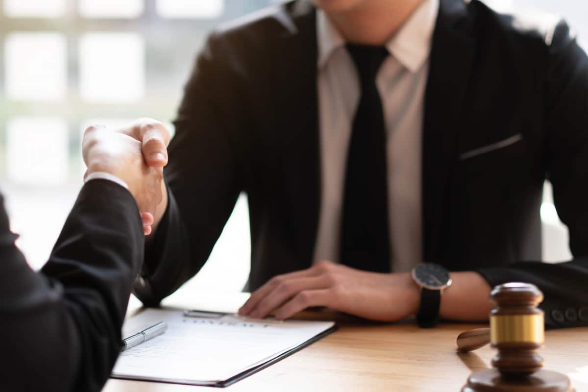 Man dressed in suit shaking hands with another person across a table. There is paper and a pen between them and there is also a gavel on the table.
