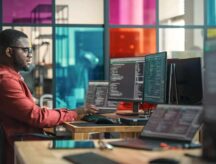 Man Writing Lines Of Code On Desktop Computer With Multiple Monitors and Laptop in Creative Office