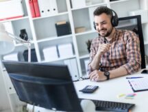 A bearded man in a plaid shirt and dark trousers works in a bright office