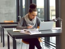 A young woman sits at a table with books and a laptop.