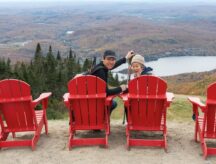 Two people sit atop a mountain view overlooking a river and small forrest below.