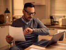 A man with glasses sitting at a table with a laptop, holding a piece of paper and checking the time on his watch.
