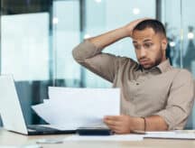 A stressed man sitting at a table holding up one hand to his head in exasperation while holding up a number of papers in the other.