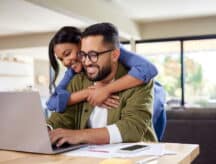 Man sitting at a table using a laptop, while a woman embraces him from behind—both are smiling.