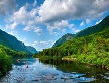 A group of kayakers row down a wide river on a beautiful summer day, with mountains in the background.