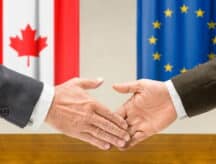 The Canadian flag and European Union flag draped against a wall, as representatives of both shake hands in the foreground (only hands visible).