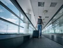 A man with a backpack and a luggage in hand at an airport terminal using a moving walkway.