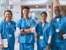 Four nurses in scrubs standing in a hospital, smiling, with stethoscopes around their necks.