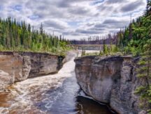 The Sambaa Deh Falls Territorial Park in the Northwest Territories on a partially sunny day, with the falls and highway bridge visible in the back.