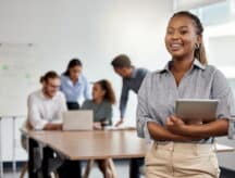 Young woman dressed in office attire holding a tablet, with a group of four workers behind her.