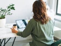 A woman sitting at a table while using a laptop and holding a coffee, shot from behind.