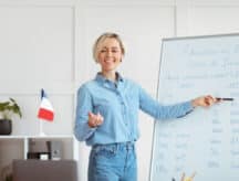 Blonde woman standing by and point at a whiteboard which has French writing on it.