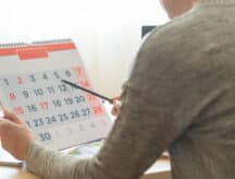 Person in grey long-sleeve shirt seated at a desk holding a calendar in one hand and a pen in the other, which they are using to point at the calendar.