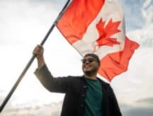 A man standing outside, proudly holding up a large Canadian flag, smiling as it waves in the wind.