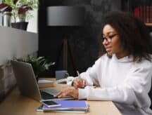Black female student working on a laptop, at a desk, with a pen in hand.