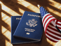 Two passports, the top passport clearly a U.S. passport, lying on a table beside a small American flag.